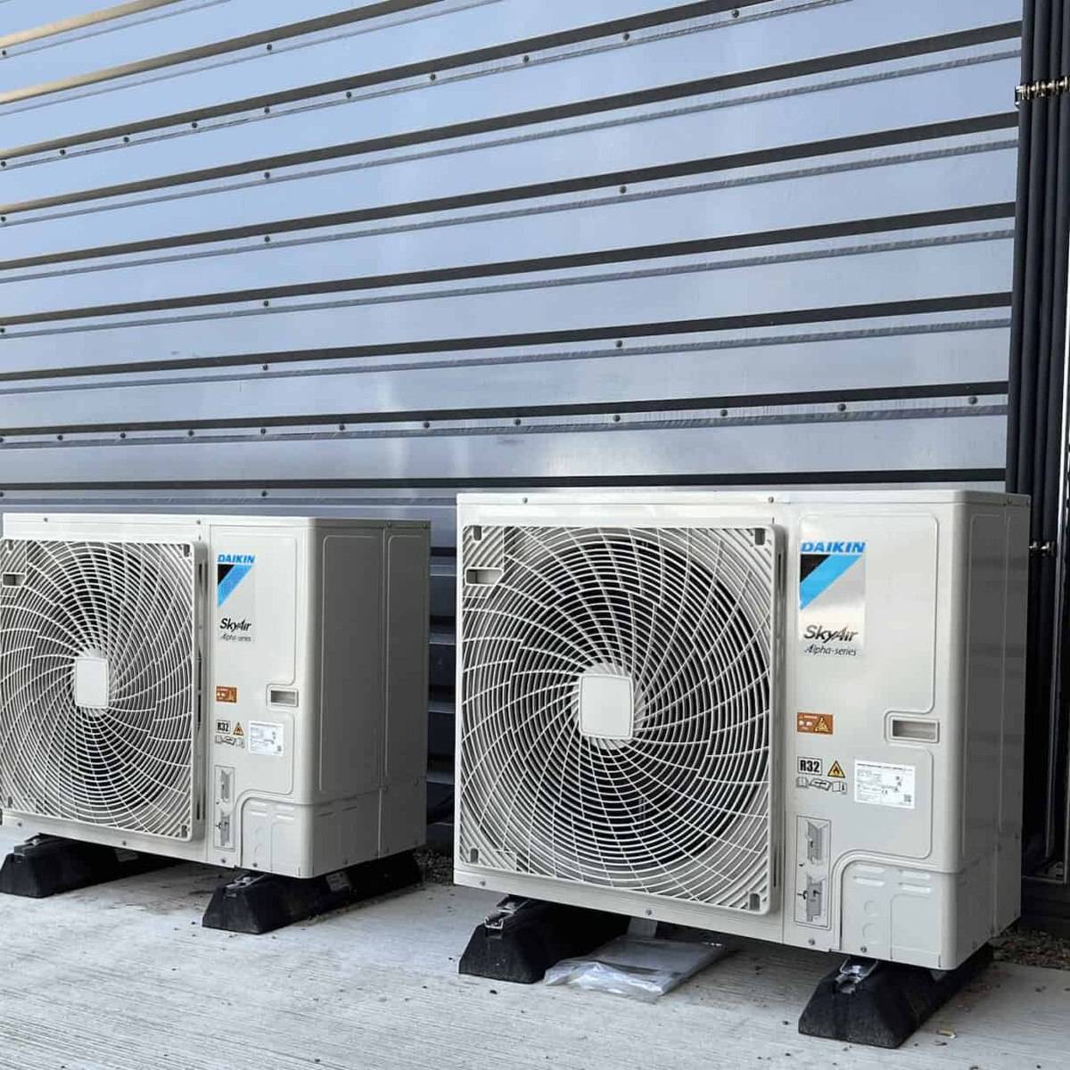 Two Daikin Sky Air outdoor units standing on anti-vibration feet against the clad wall of a commercial building, with the pipework dropping down behind them