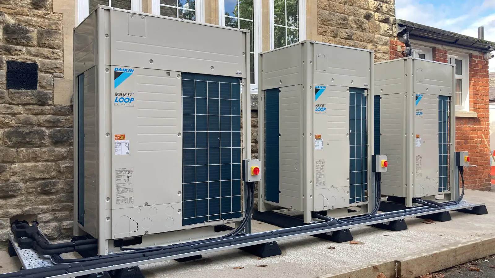 Three Daikin VRV outdoor units standing side by side on a steel frame against a stone building, each with its own isolator, and the pipework run in tray along the base